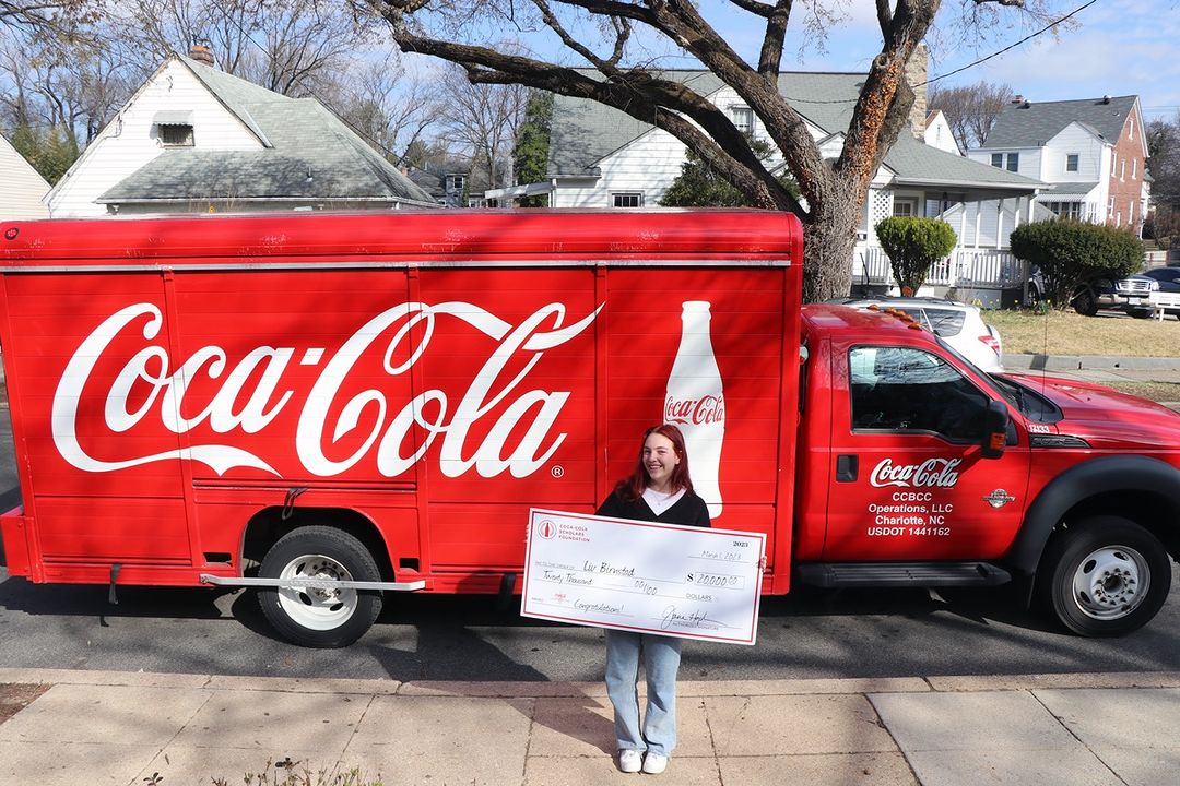 Liv is smiling in front of a Coca-Cola truck, holding her scholarship cheque