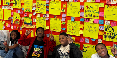 post it notes pinned across a wall with students sitting beneath it waving at the camera