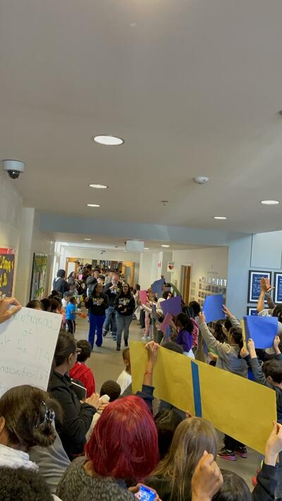 students and parents line the walls of a school hallway, all holding celebratory posters, while the graduating seniors march through